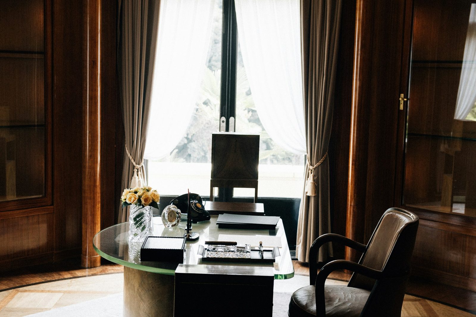 Quiet-luxury home study with warm walnut-panelled walls, sheer white curtains over a tall window, a round glass-top desk with yellow roses, and a leather armchair