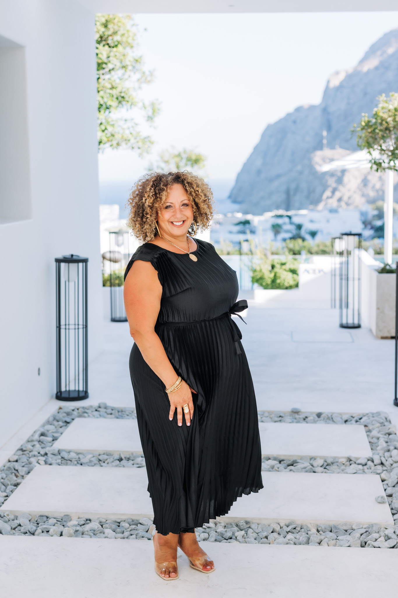 Melina Bradley standing outdoors in a black dress with Santorini Greece scenery in the back ground