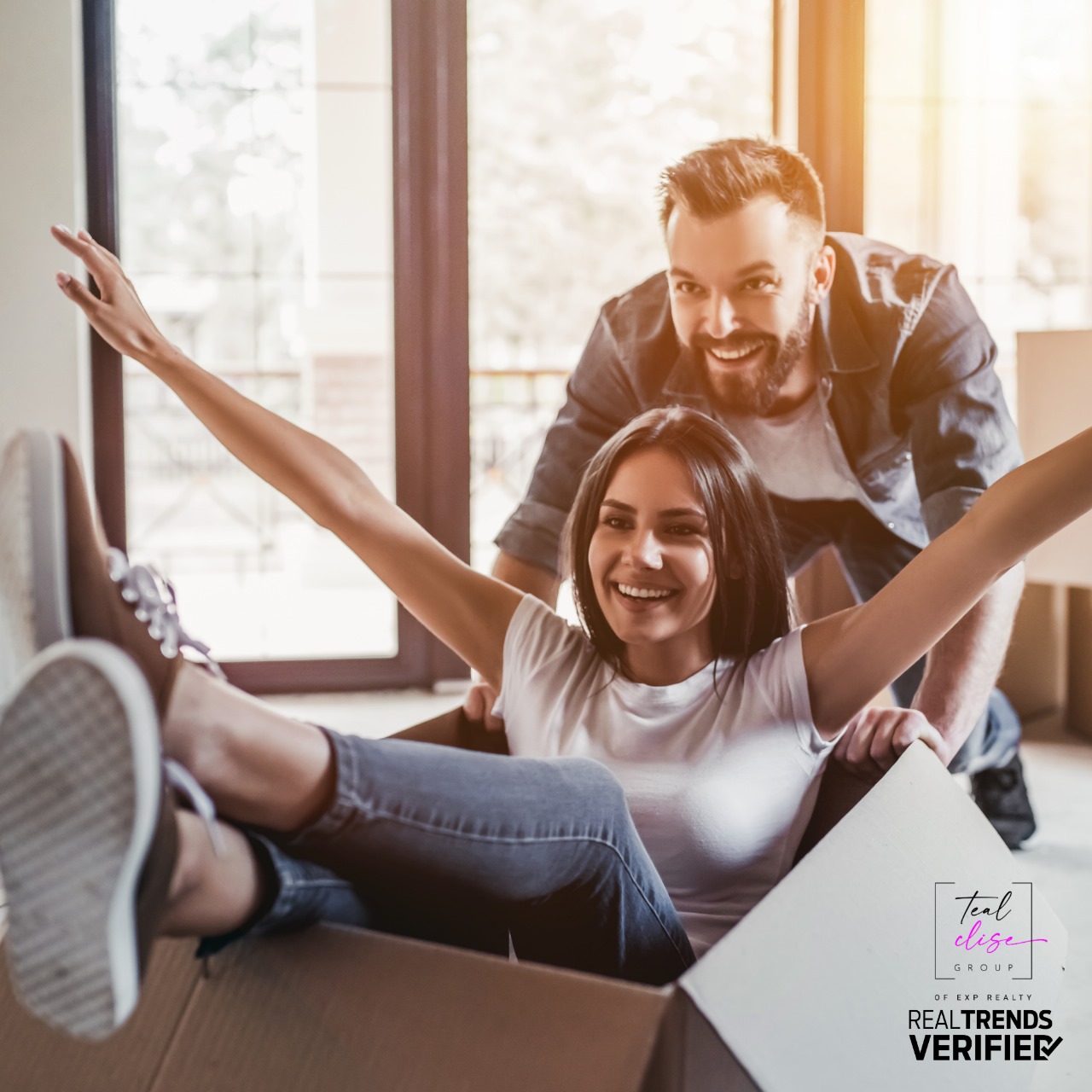 A smiling couple celebrates moving into their new home. The woman sits playfully in a cardboard box while the man pushes her, capturing the excitement of a smooth home transition. Image reflects the emotional reward of using a Home of Choice Contingency when selling and buying in Maryland.