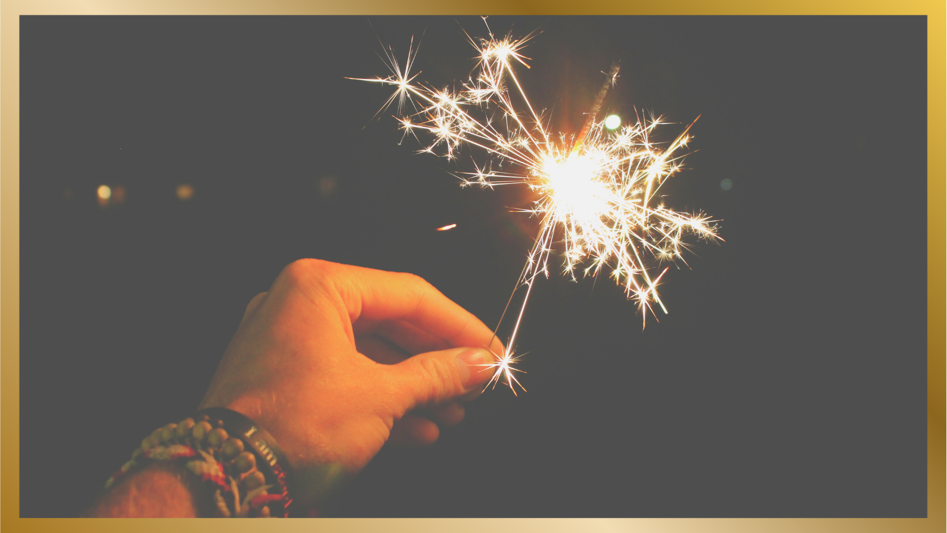 Hand with stacked beaded bracelets holding a sparkler against a dark night sky