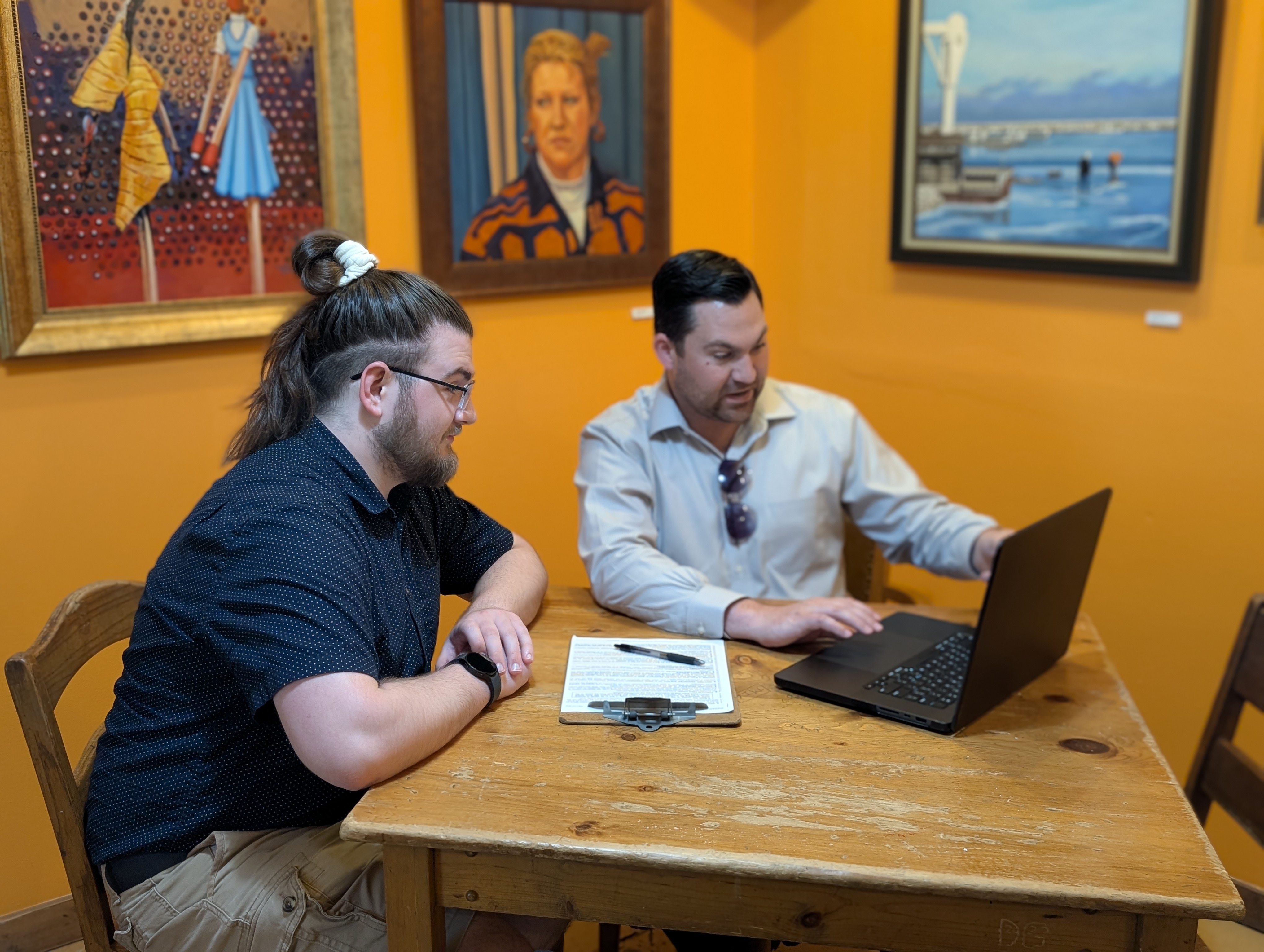 Two men sitting in front of a computer at a coffee shop. One pointing to the screen and one looking at it carefully