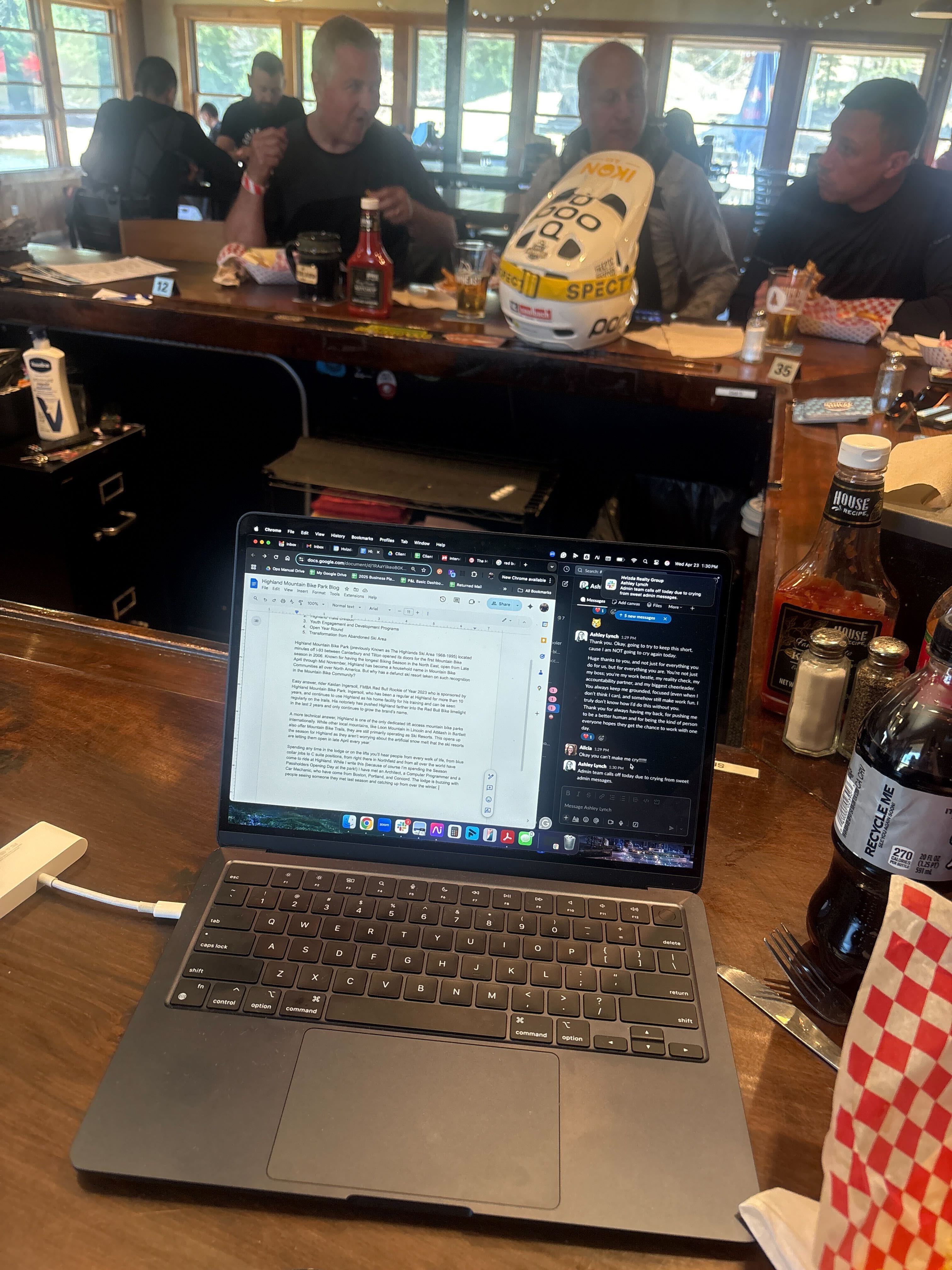 Laptop sitting on top of a wooden bar top. In the back sits riders behind the bar enjoying food and drinks while a helmet sits on top of the bar in front of them.  
