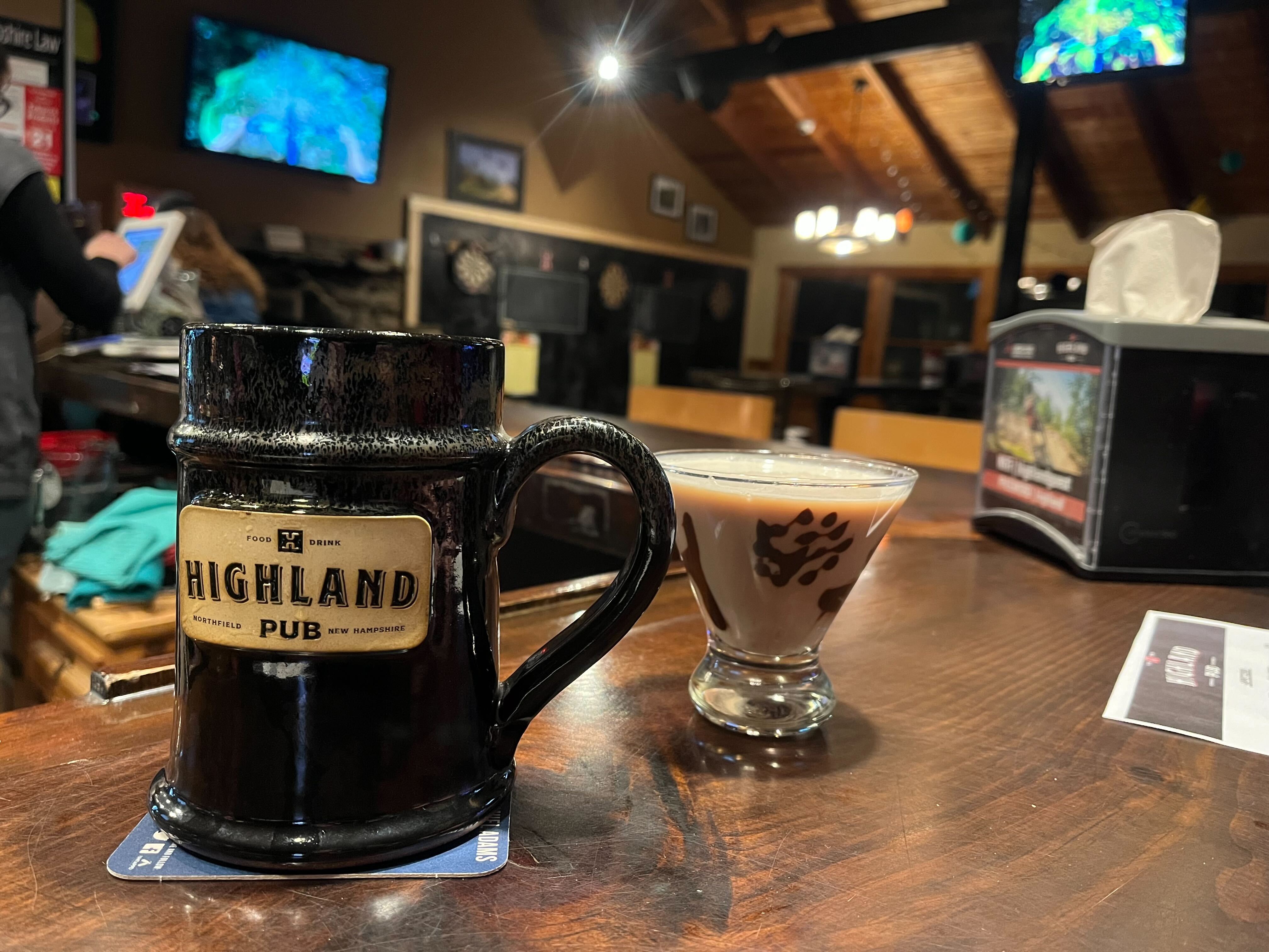 A handcraft mug with a highland pub logo on the front sits on the bar next to a martini glass