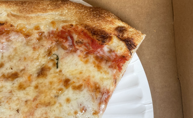 Close-up of the corner of a large slice of cheese pizza, on a white paper plate inside of a pizza box