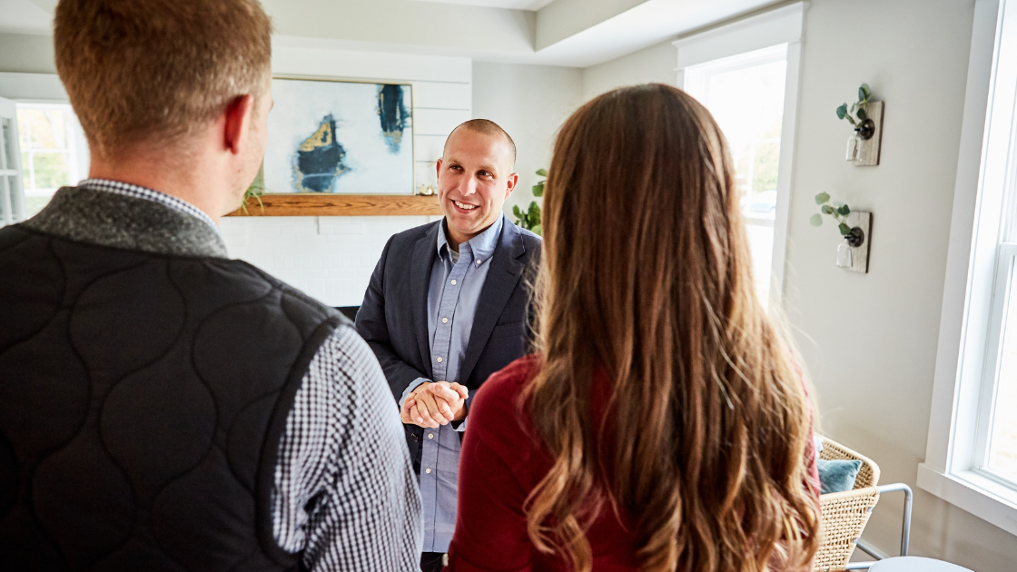 A real estate agent faces the camera as he warmly greets a homebuying couple at a house
