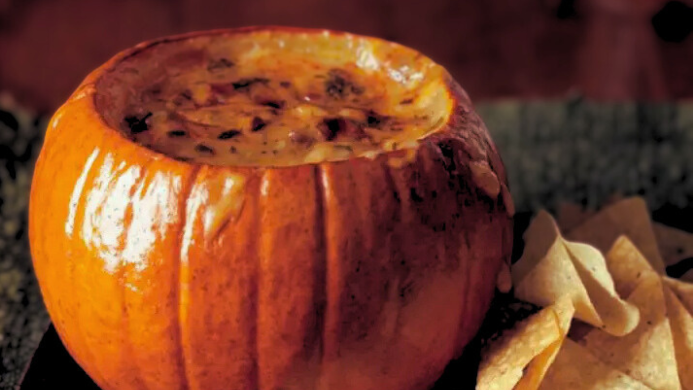 A small orange pumpkin has its top cut off. Inside, toasted melty cheese fills the pumpkin, spilling from the brim. The right of the pumpkin, a pile of tortilla chips