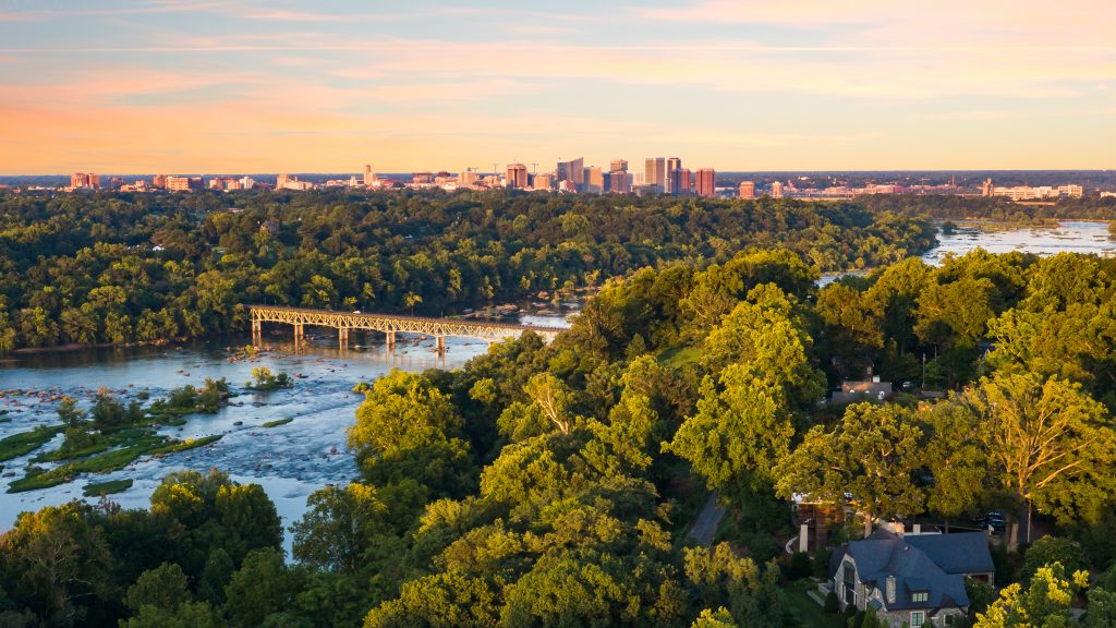 An aerial photograph featuring the james river in Richmond, VA. Across the river a concrete bridge stretches from one end to the other. In the distance, the Richmond City skyline rises just above the horizon. 