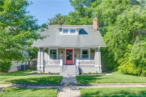 A 1930s bungalow with steeped roof and upstairs bump-out sits central in the photo. In the foreground, a sidewalk runs along the length of the photo. Surround hte house is grass as well as tall leafy trees and foliage