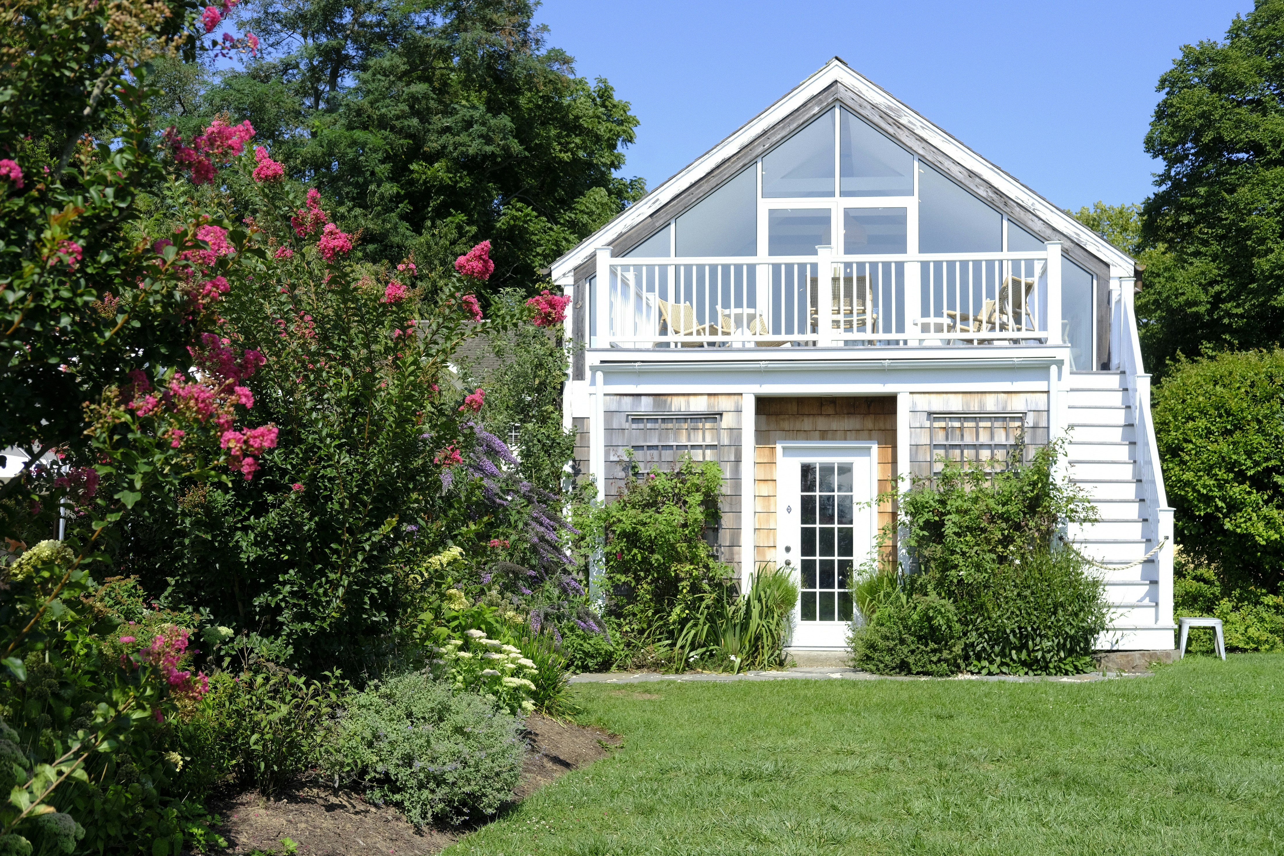 Detached backyard accessory dwelling unit with white siding, large windows, an upper balcony, and a landscaped garden.
