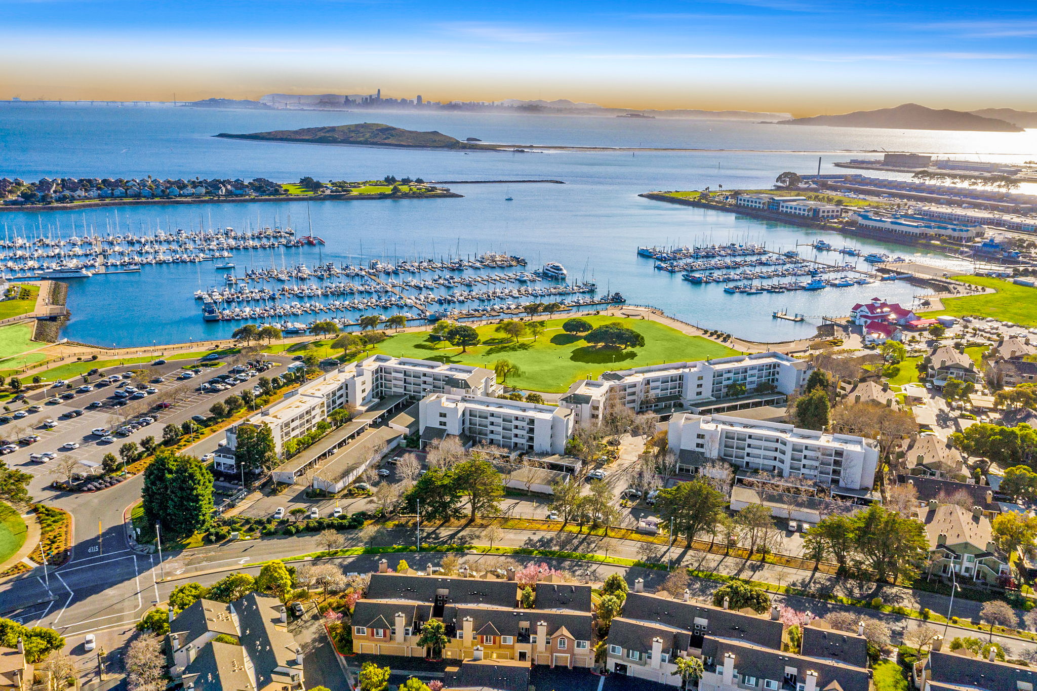 Aerial view of Marina Bay, Richmond, showing waterfront condo buildings, a marina filled with sailboats, green park space, the Bay, and the distant San Francisco skyline.