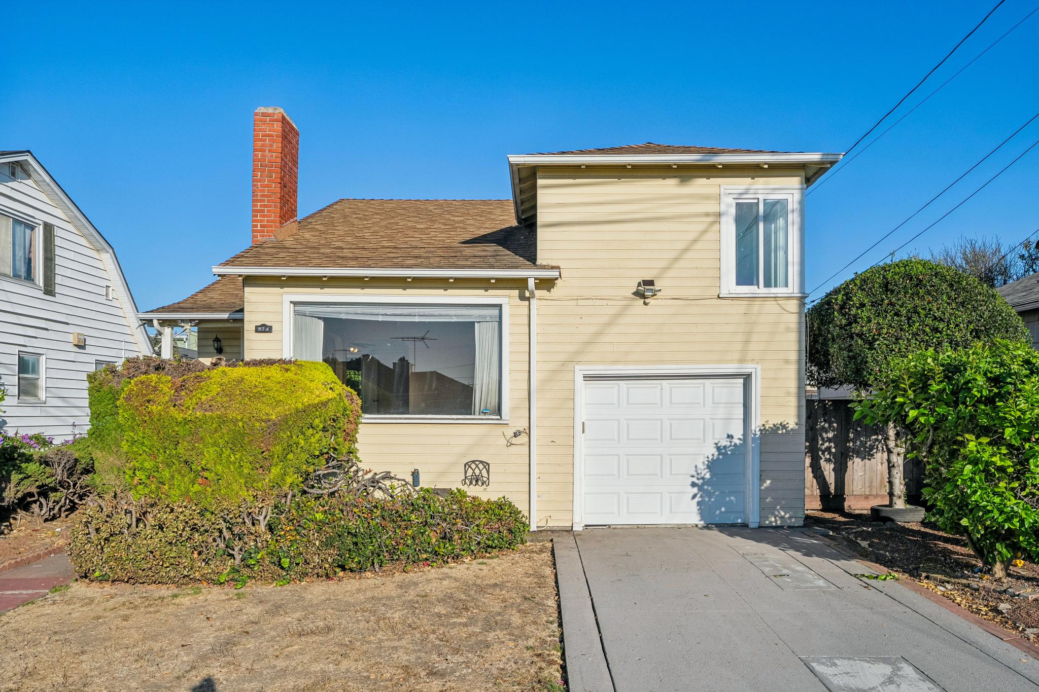 Front exterior of 974 33rd St in Richmond, CA, with beige siding, a brick chimney, attached garage, driveway and front hedges.