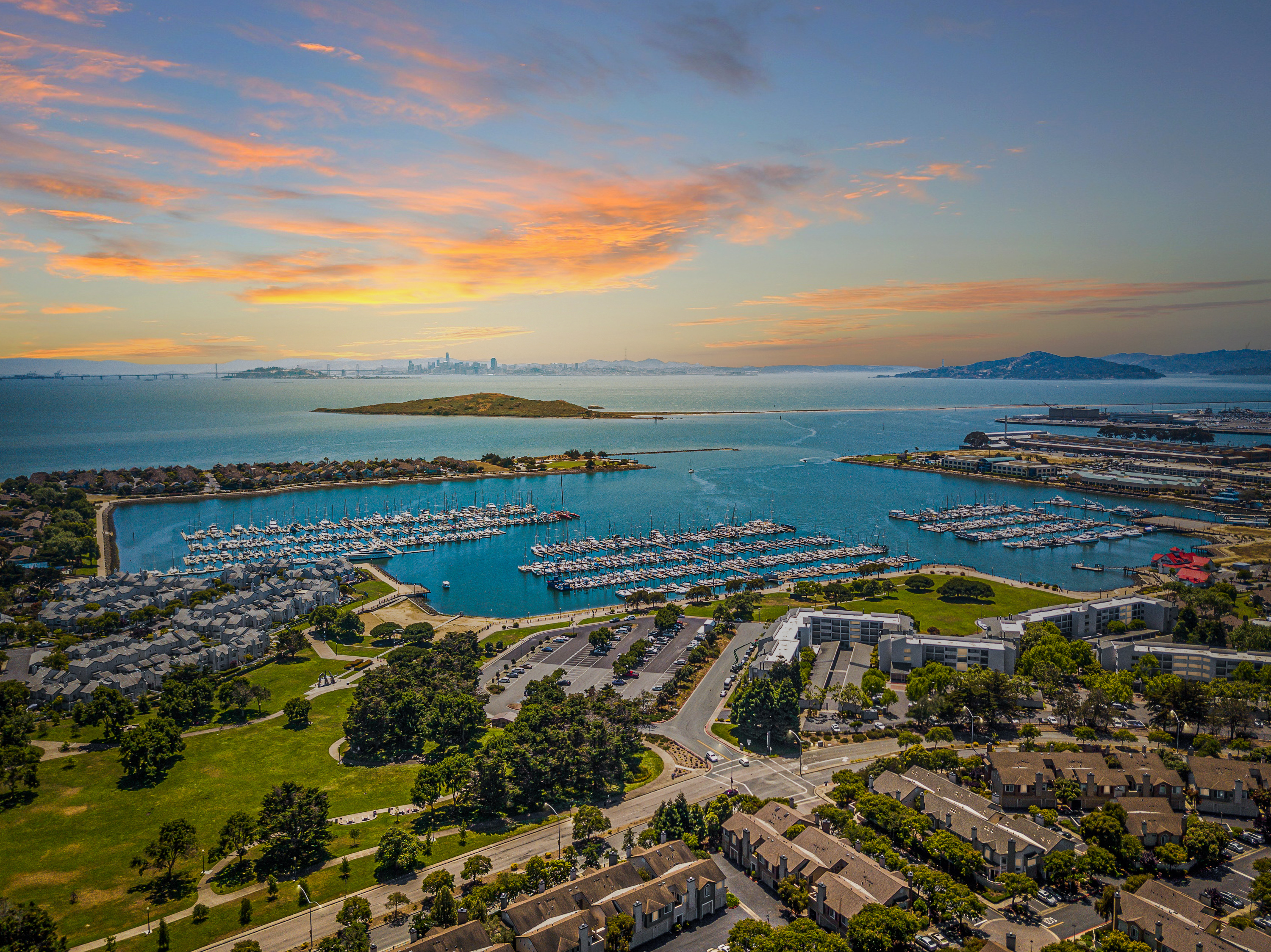 Aerial view of Marina Bay in Richmond, California, with sailboat docks, waterfront homes, green spaces, and San Francisco Bay beneath a colorful sunset sky.