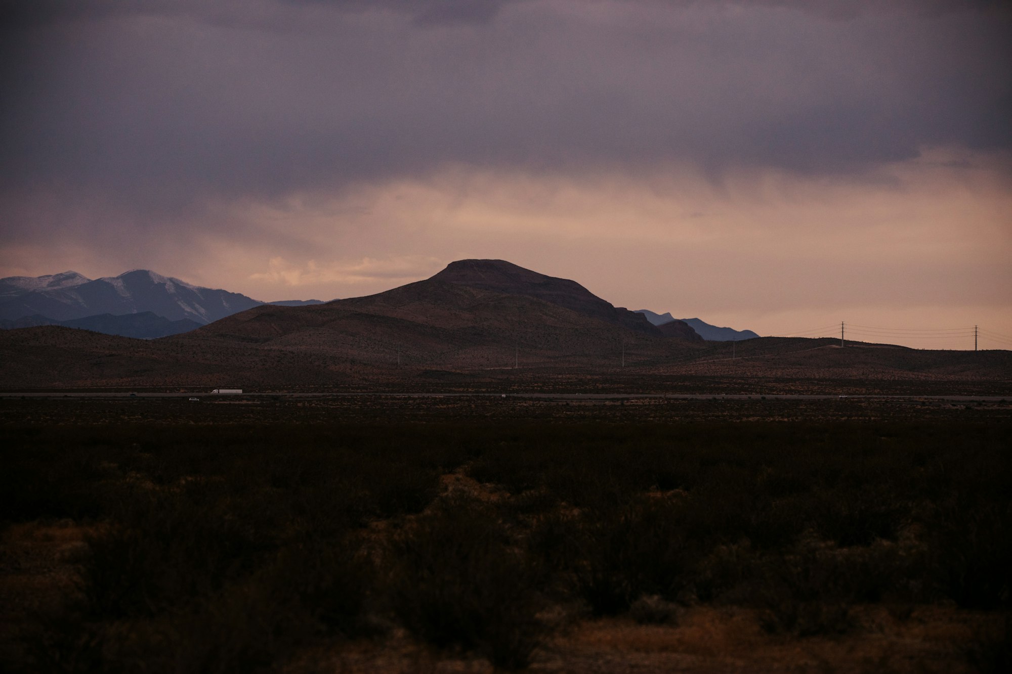 Desert scrub in the foreground with a brown hill and snow-dusted mountains under a cloudy, moody sunset sky.