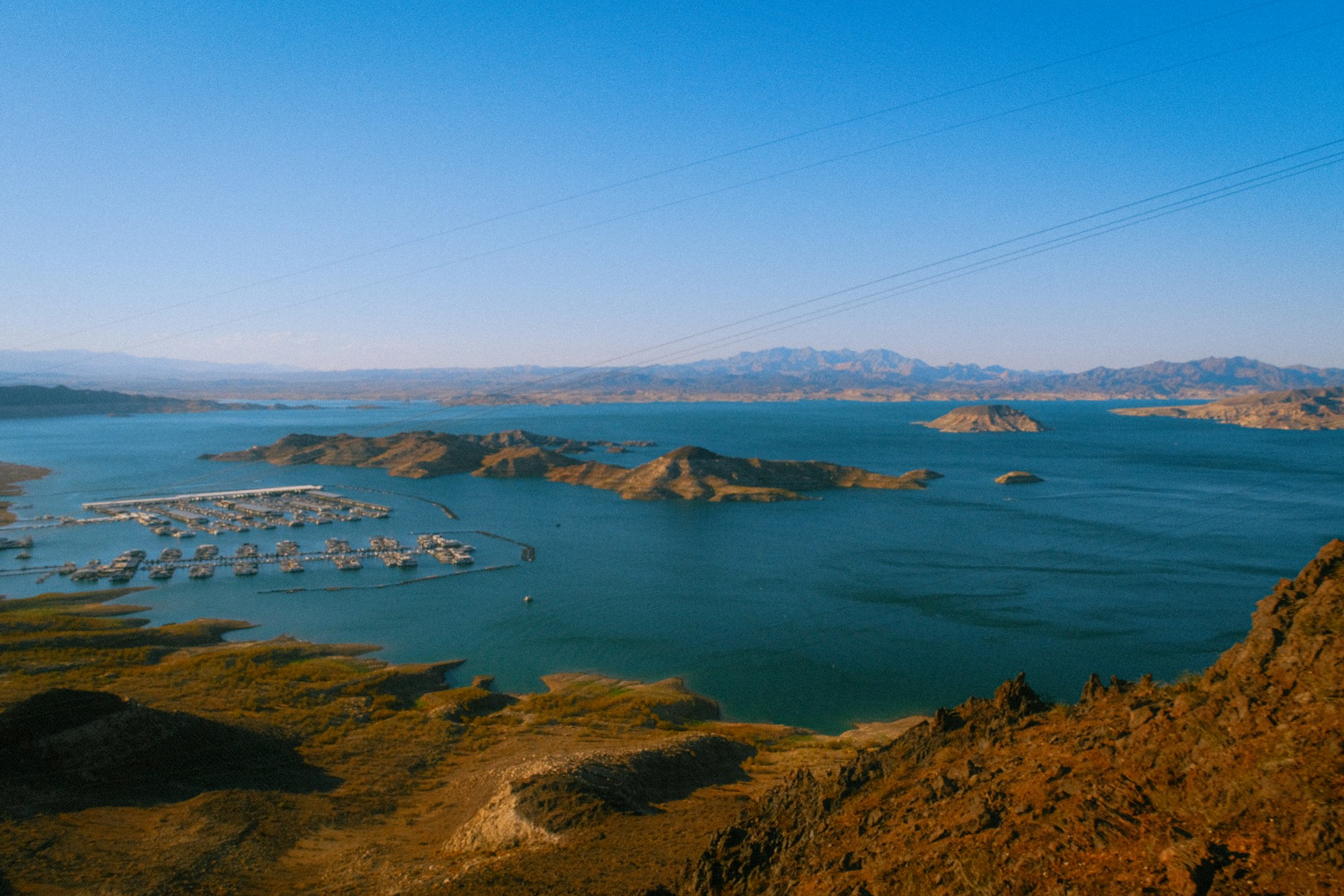 Blue lake water with rocky desert islands, a small boat marina, and distant mountains under a clear sky.