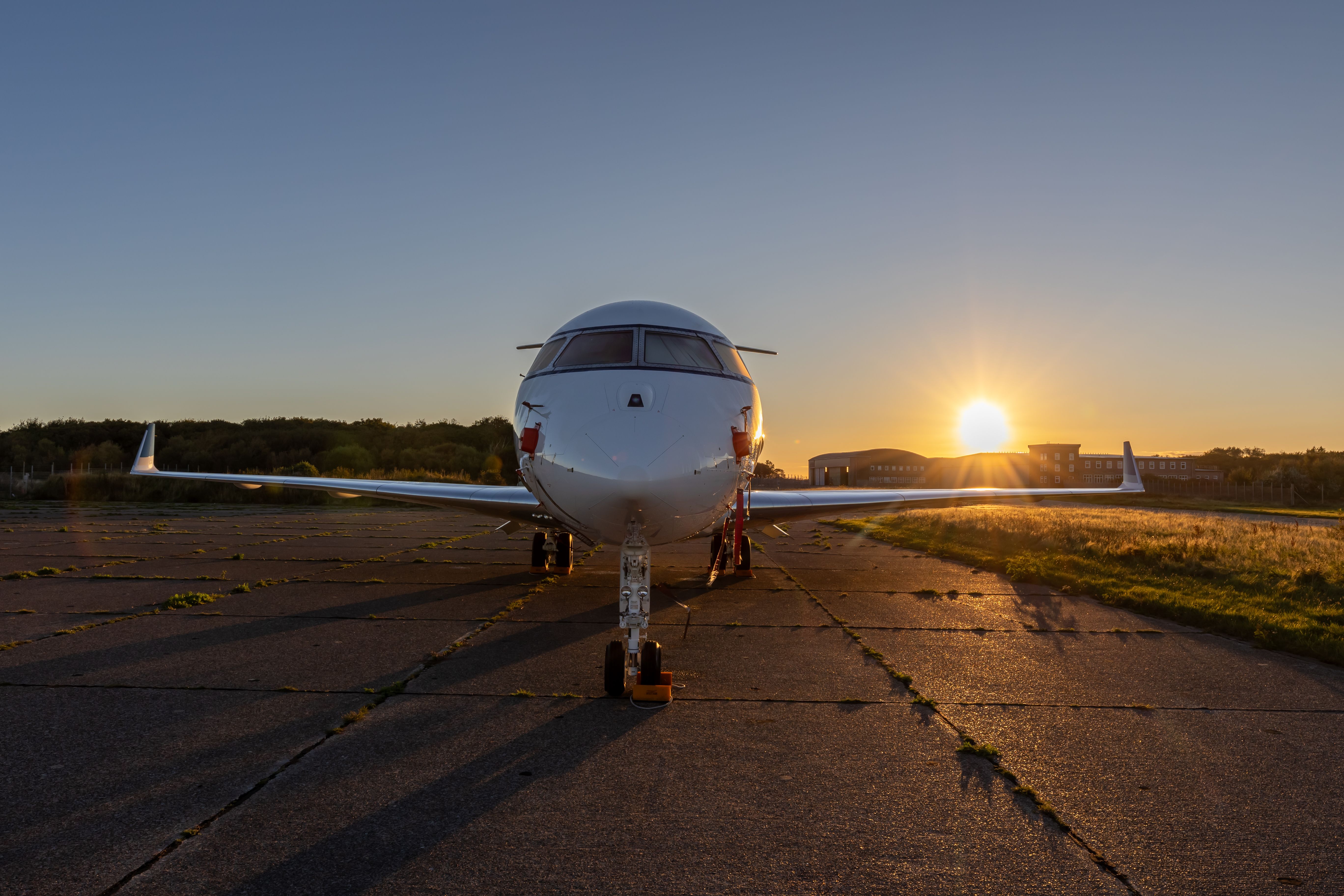 White business jet parked head-on on an airport ramp at golden hour, with the low sun flaring beside the nose and long shadows across the tarmac
