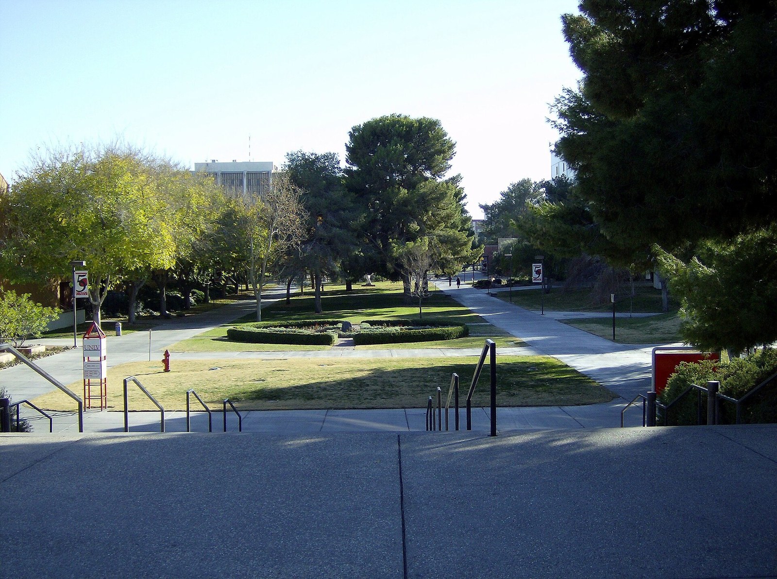 Tree-lined central quad on the University of Nevada, Las Vegas campus on a clear day, viewed from concrete steps toward a grassy lawn, walkways and a campus building.