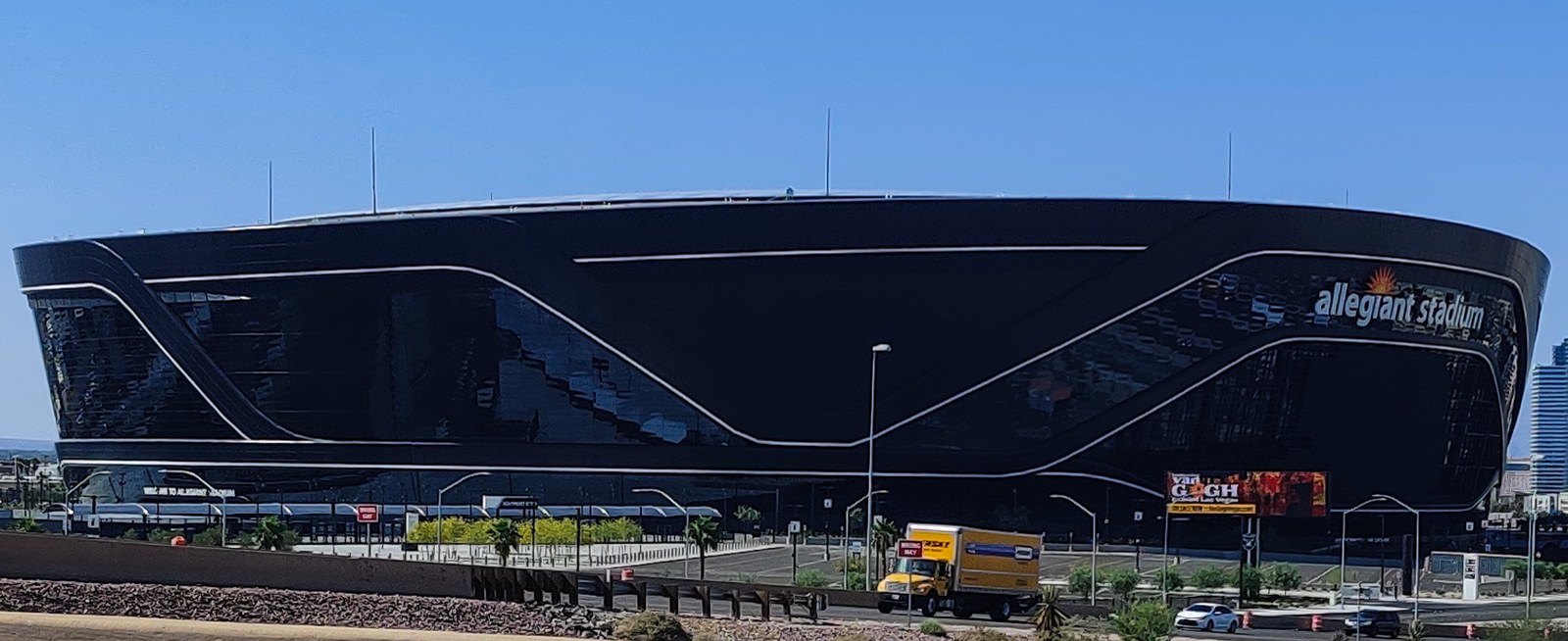 Daytime exterior of Allegiant Stadium in Las Vegas, a curved black-glass facade with white sweeping lines and the 'allegiant stadium' sign, under a clear blue sky with palm trees and a yellow box truck in the foreground.