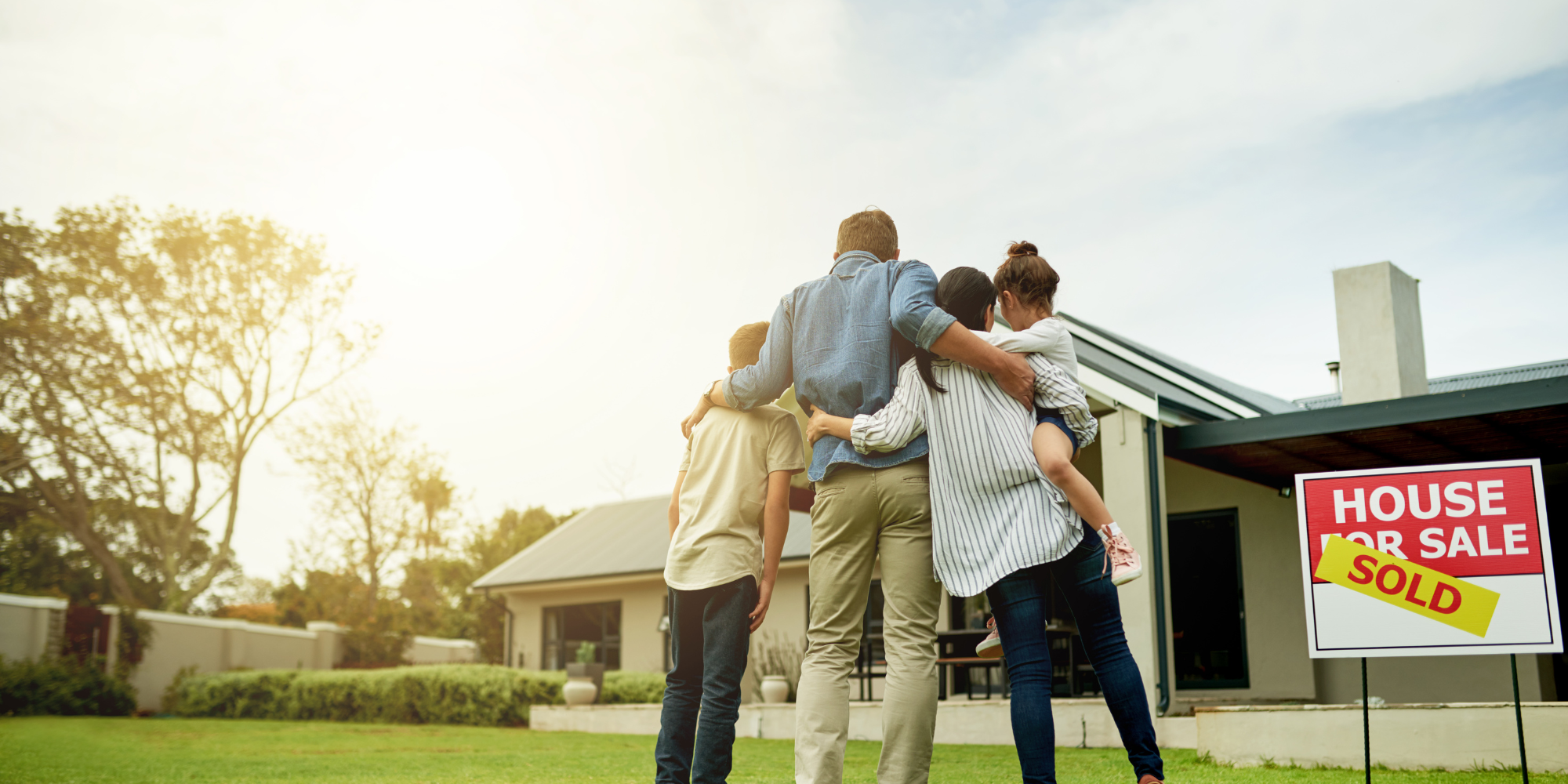 Happy family standing in front of their new home with arms around each other, looking at the house with a “Sold” sign in the yard, symbolizing a successful home purchase.