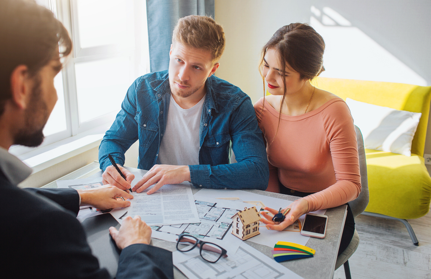 Young couple discussing home purchase with real estate agent, reviewing documents and house plans at a modern office table.