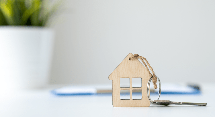 A small wooden house-shaped keychain with silver keys resting on a white surface, representing home ownership and the successful completion of a real estate transaction.