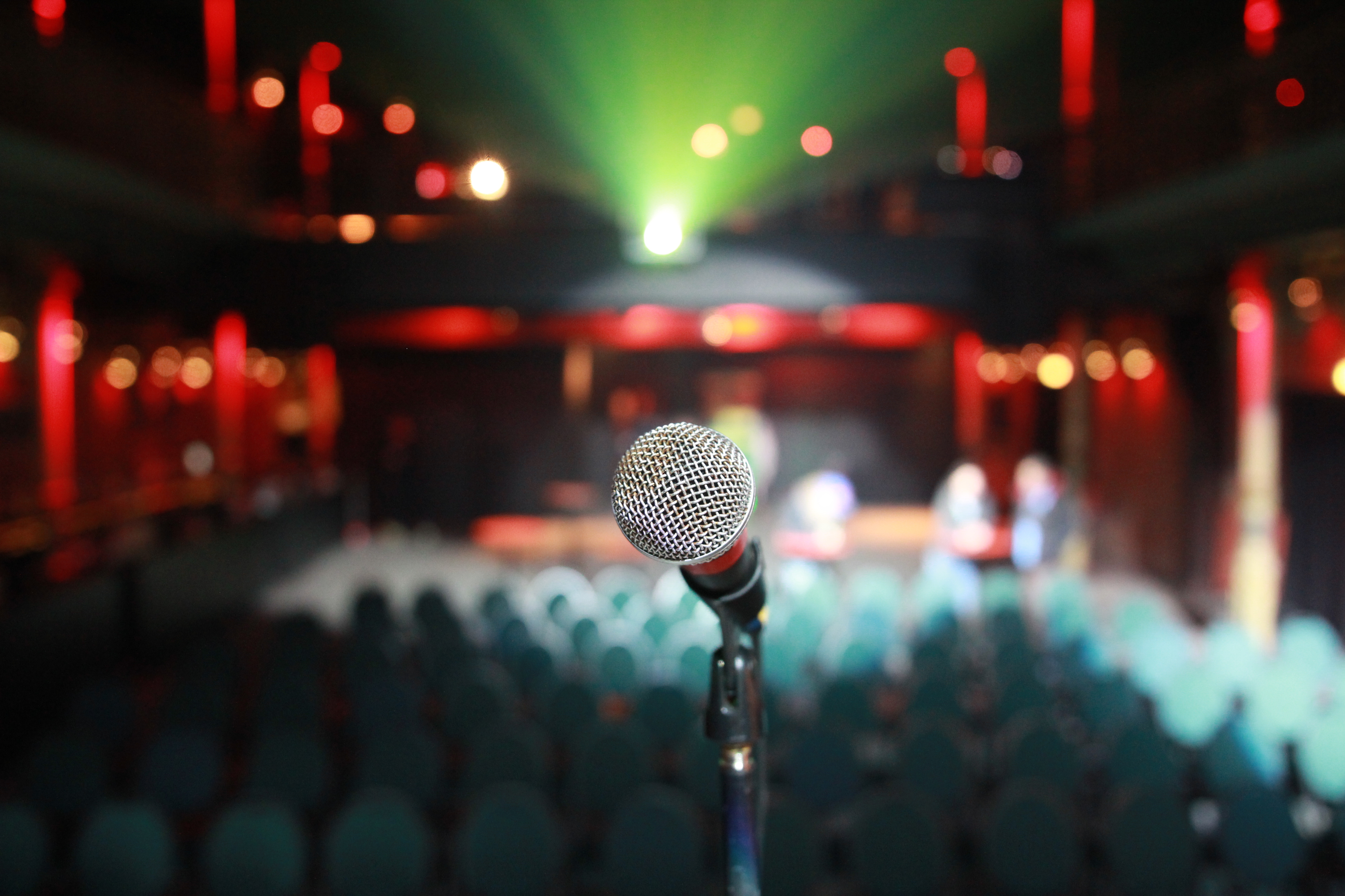 Microphone on a stand facing rows of seats in a dimly lit concert hall with warm red and green stage lights.