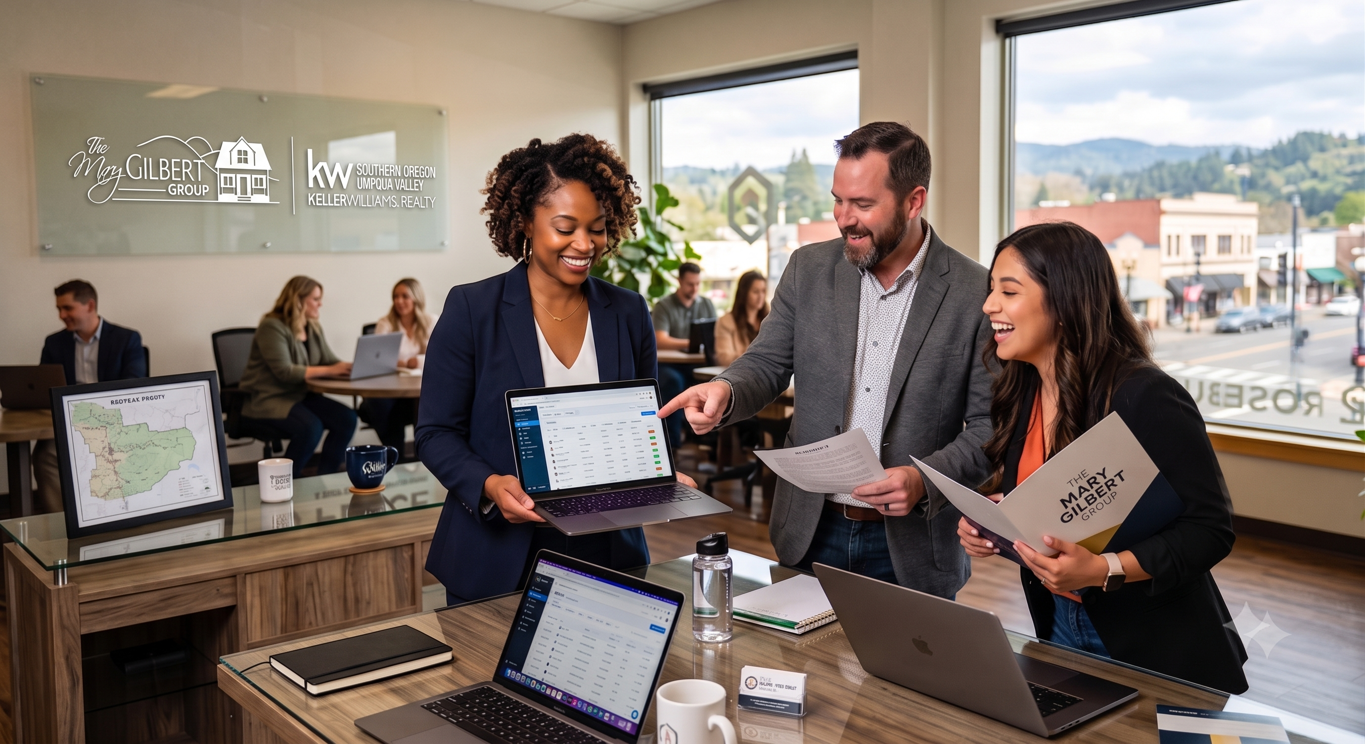 Three diverse real estate agents smiling and collaborating over a laptop and paperwork inside a modern Roseburg office. The frosted glass wall in the background prominently features The Mary Gilbert Group and Powered by PLACE corporate logos.
