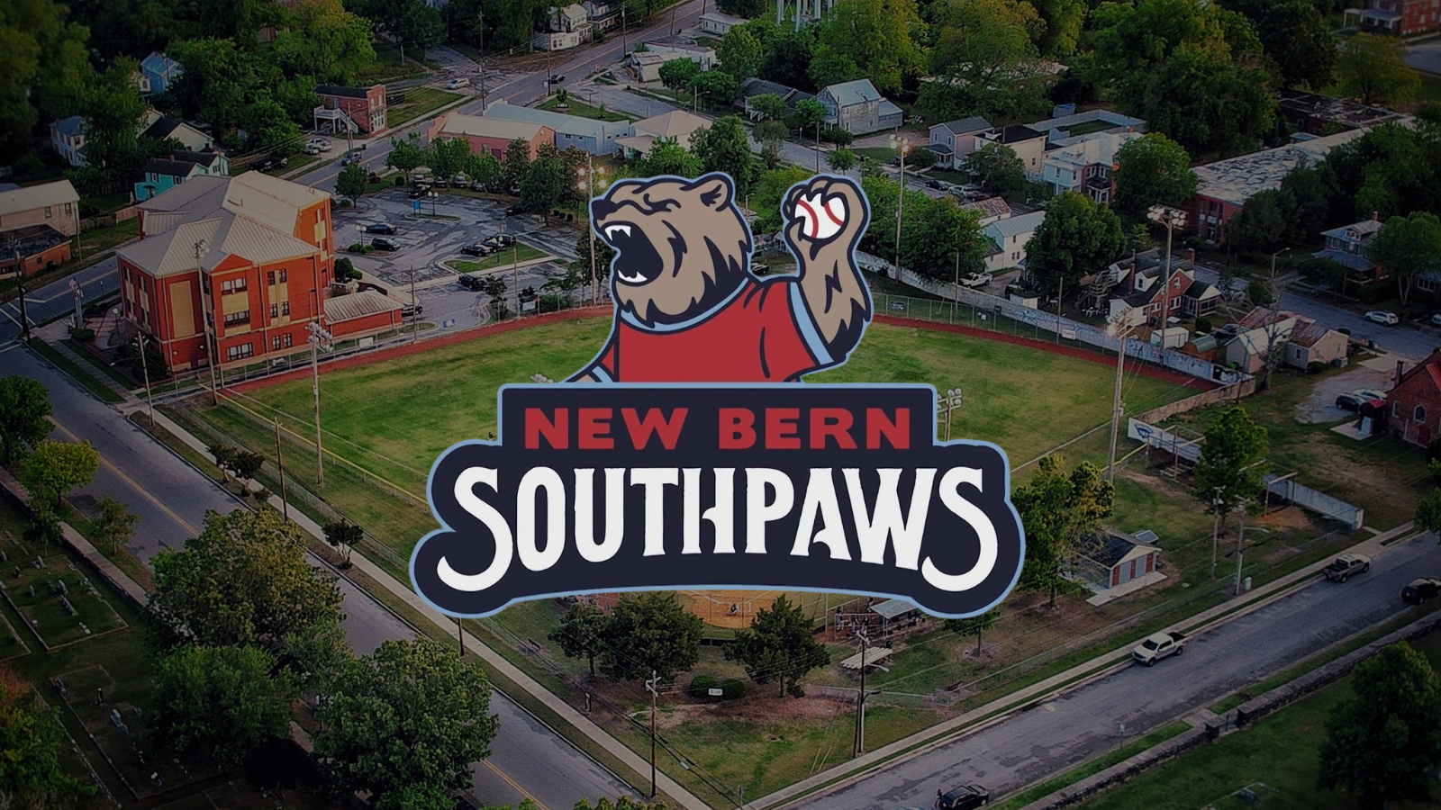 Aerial view of the New Bern Southpaws baseball stadium with the official Southpaws logo—a roaring bear holding a baseball—centered over the field. Surrounded by historic New Bern neighborhoods and tree-lined streets.