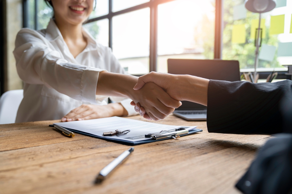 Two people shake hands across a desk with paperwork and a clipboard, symbolizing a successful real estate transaction, contract signing, or home sale agreement.