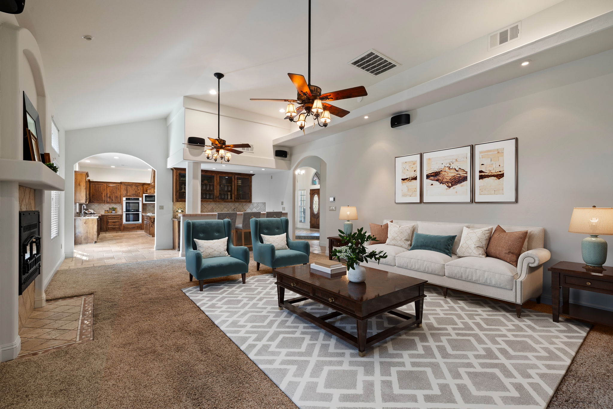 Staged open-concept living room in a Bakersfield home featuring vaulted ceilings, carpeted floors, modern lighting, a fireplace, and a spacious kitchen beyond the archway.