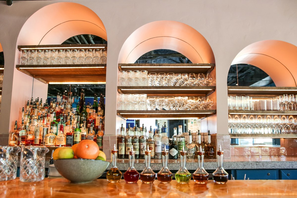 Restaurant bar with three arched, backlit shelves of liquor bottles and glassware above an onyx counter lined with small bitters bottles, crystal pitchers and a bowl of fruit