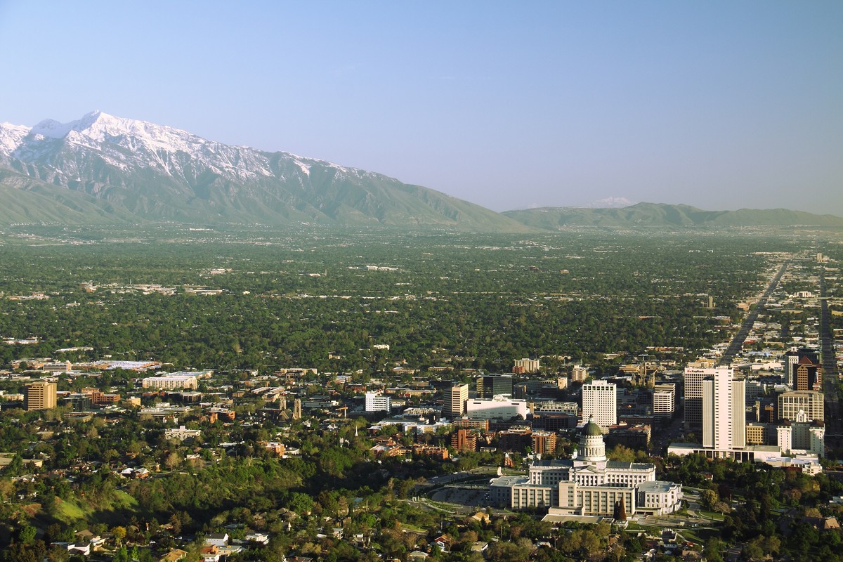 Utah State Capitol and downtown Salt Lake City seen from Ensign Peak, with the green Salt Lake Valley and the snow-capped Wasatch Mountains behind under a clear evening sky.