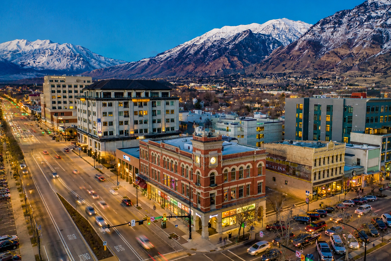 Aerial view of downtown Provo, Utah at dusk showing a mix of historic brick architecture and modern buildings along a central street corridor, with illuminated storefronts, light traffic, and pedestrians below. Snow-capped Wasatch Mountains rise dramatically in the background under a deep blue evening sky, emphasizing the city’s valley setting and structured urban layout.
