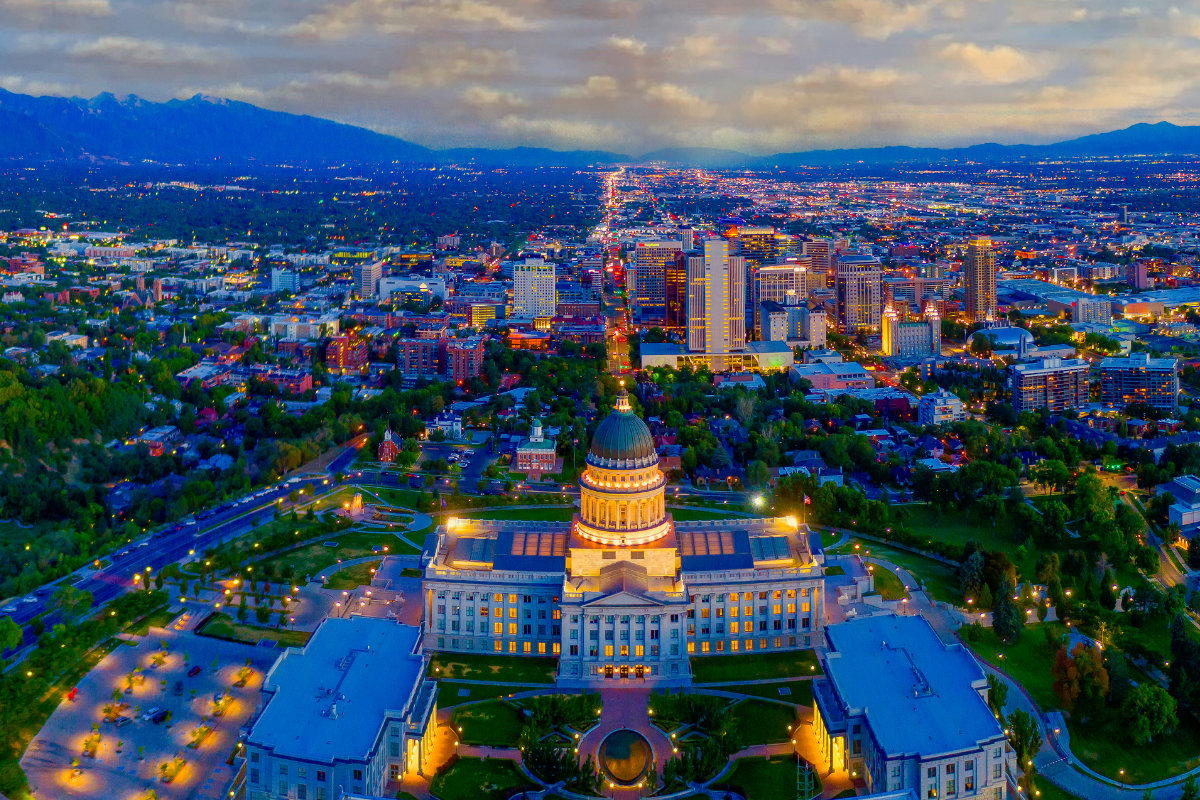 Aerial twilight view of Salt Lake City with the illuminated Utah State Capitol building in the foreground and downtown skyline extending to the Wasatch Mountains, showcasing Utah's vibrant economic center against a backdrop of natural beauty.