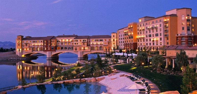 Twilight aerial view of Lake Las Vegas featuring Mediterranean-style waterfront hotels, a picturesque bridge over the lake, sandy beach area, and resort pool with lounge chairs