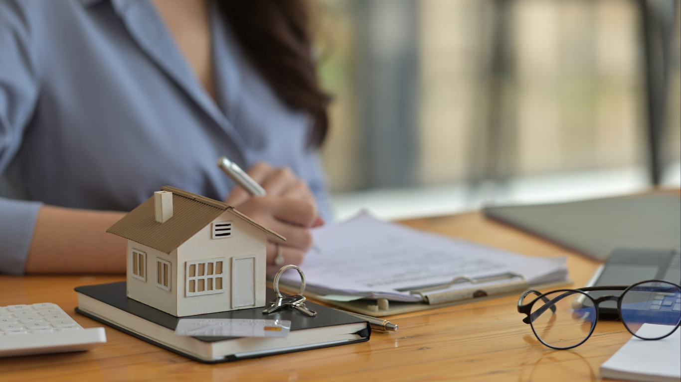 Real estate investor signing property documents with house model, keys, and calculator on desk — representing home buying, rental property investment, and refinance planning.