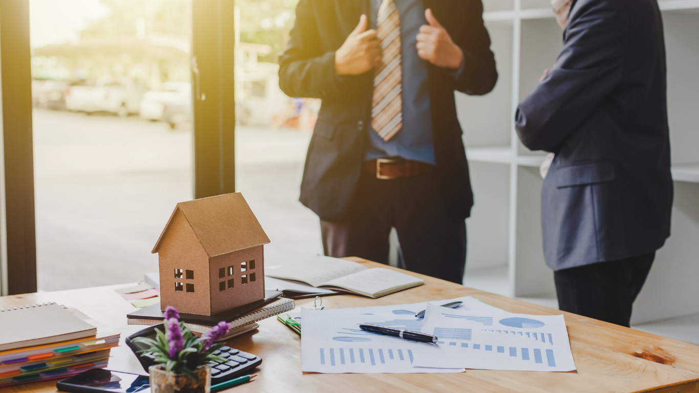 Real estate agents in suits discussing property investment strategy with a miniature house model and financial charts on the office desk.