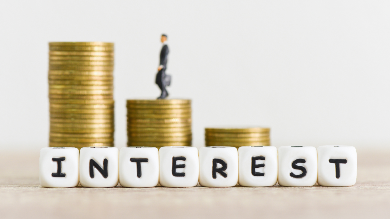 Stacks of gold coins with miniature businessman figure and word ‘interest’ spelled out in front — symbolizing high interest rates, investment growth, and real estate opportunities for investors.