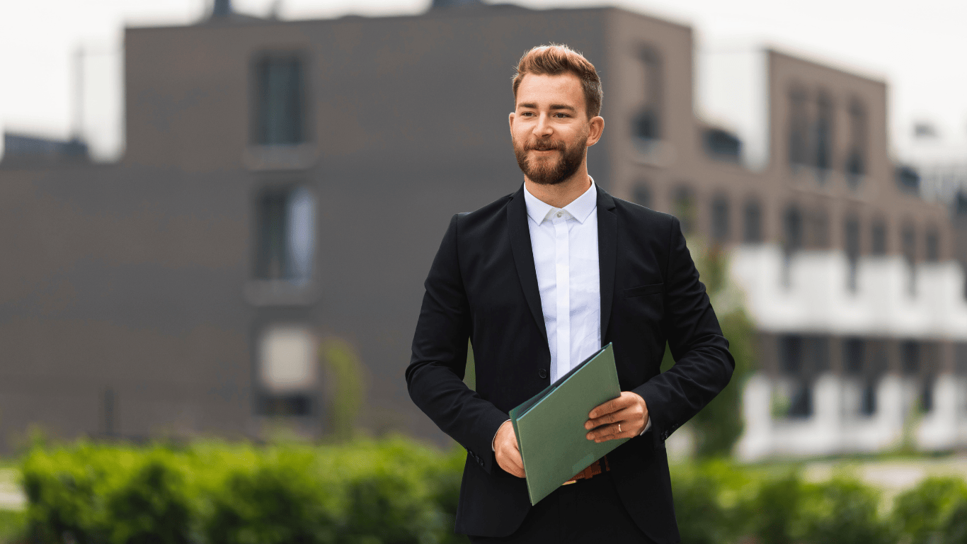 Confident real estate agent in a black suit holding a folder outside modern Los Angeles apartment buildings, representing professional property services and home selling expertise.