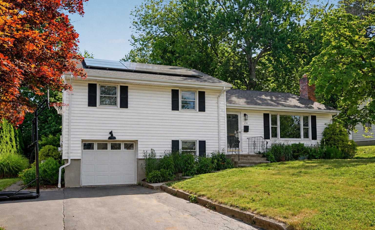 White split-level home with black shutters and an attached one-car garage, set back behind a sloping green lawn and mature trees