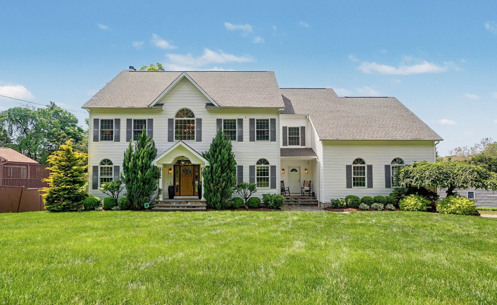 Cream center hall colonial with dark shutters, arched windows and a covered entry, set behind a deep lawn and tall evergreens
