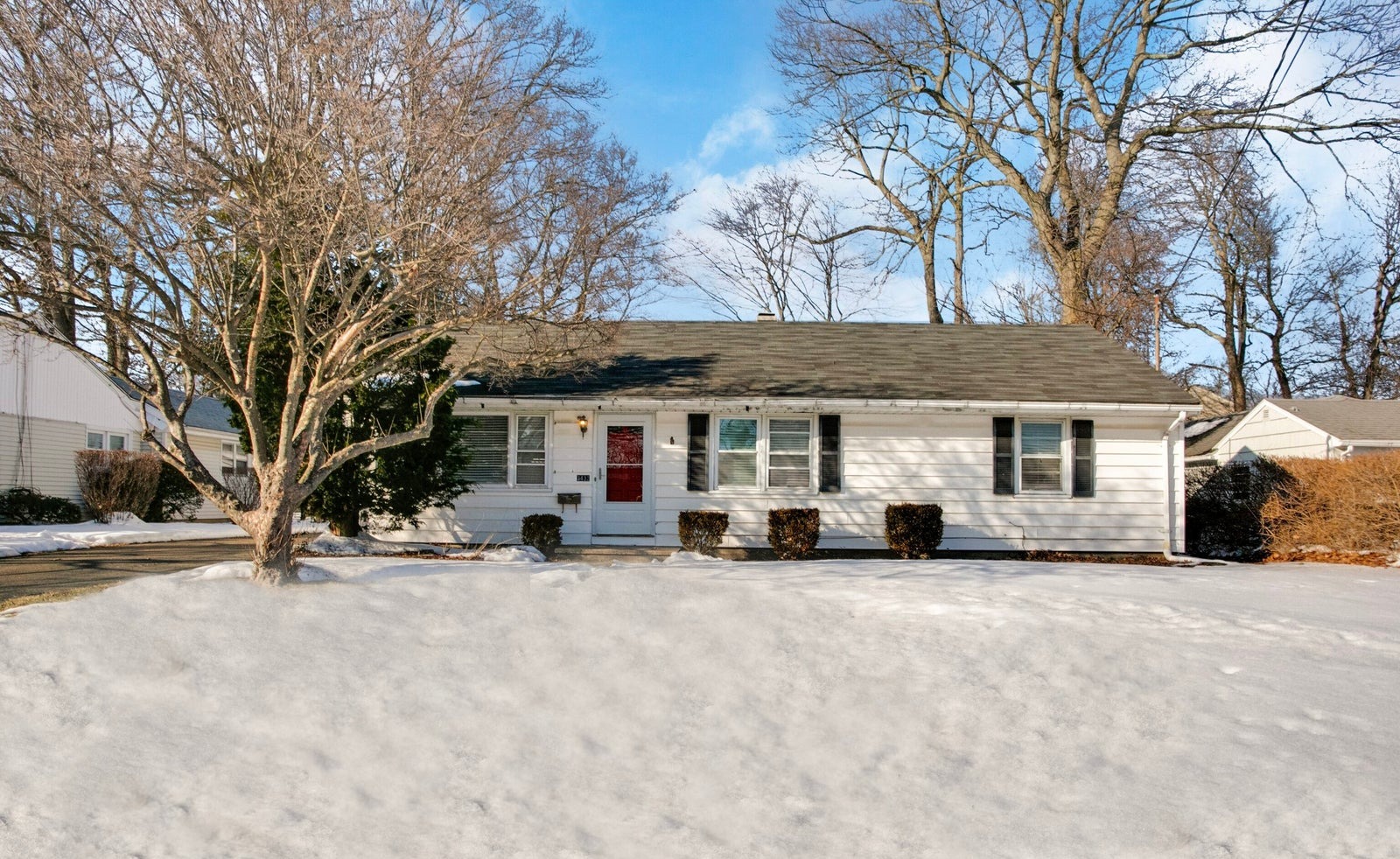 White single-story ranch house with black shutters and a red front door, set behind a snow-covered front lawn and a bare tree