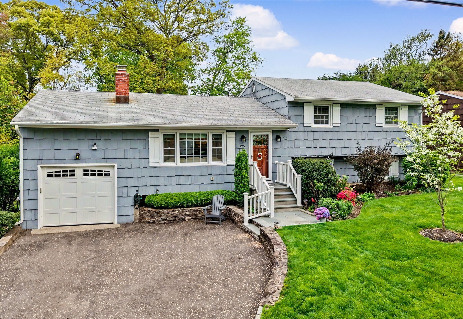 Blue-gray shingled split-level home with a white garage door, stone-edged front garden and flowering trees