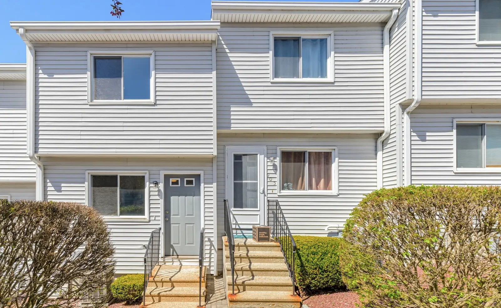 Two-story townhouse with gray vinyl siding, a gray front door, paver steps with black railings and rounded shrubs