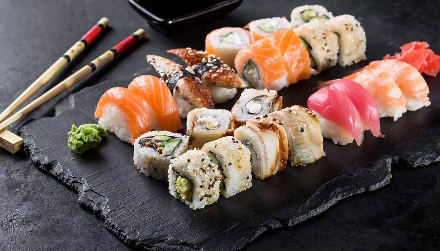 Assorted sushi and sashimi arranged on a dark slate board with chopsticks and wasabi