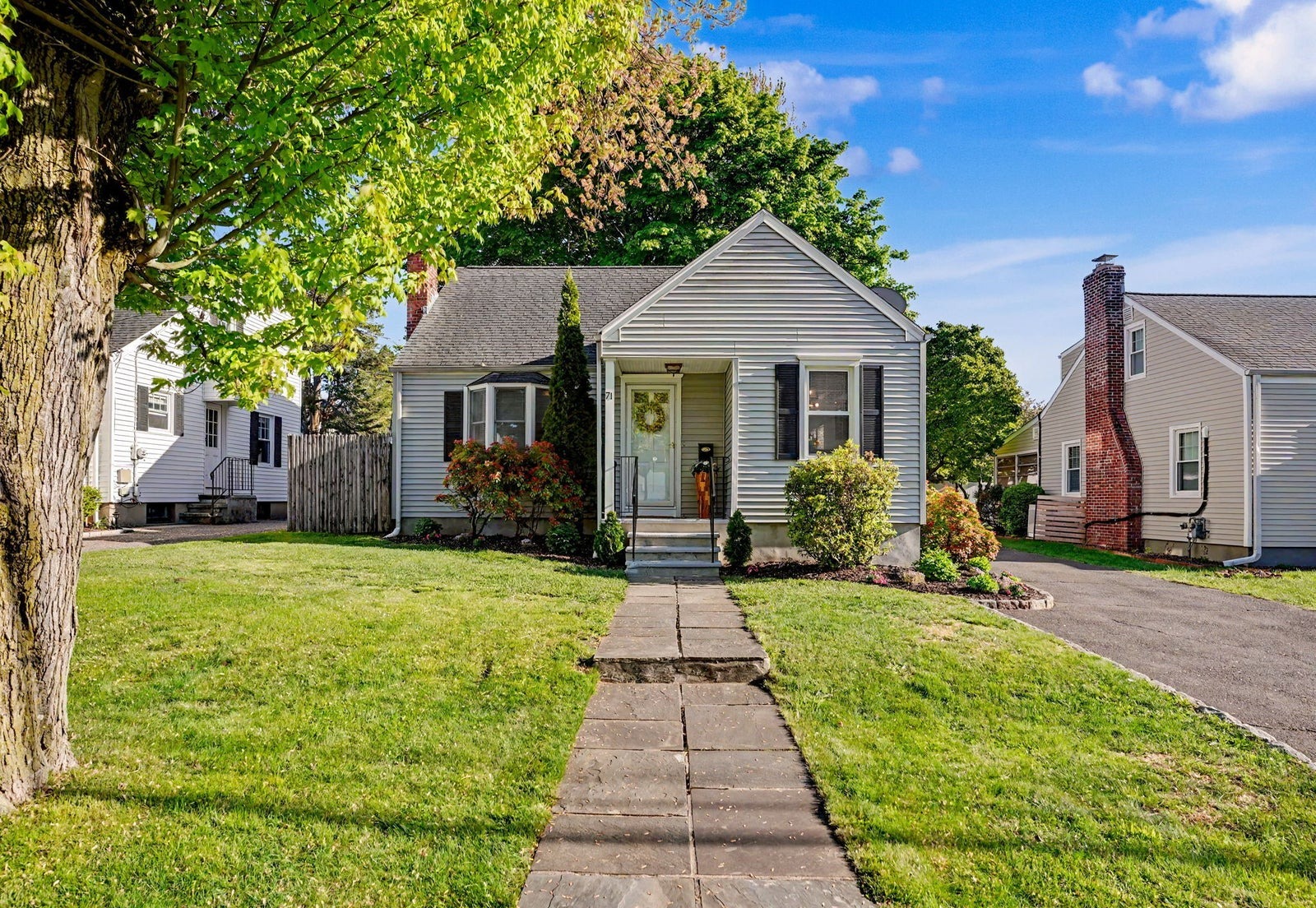 Light gray Cape Cod home with black shutters and a wreath on the front door, seen from a bluestone walkway across a green lawn
