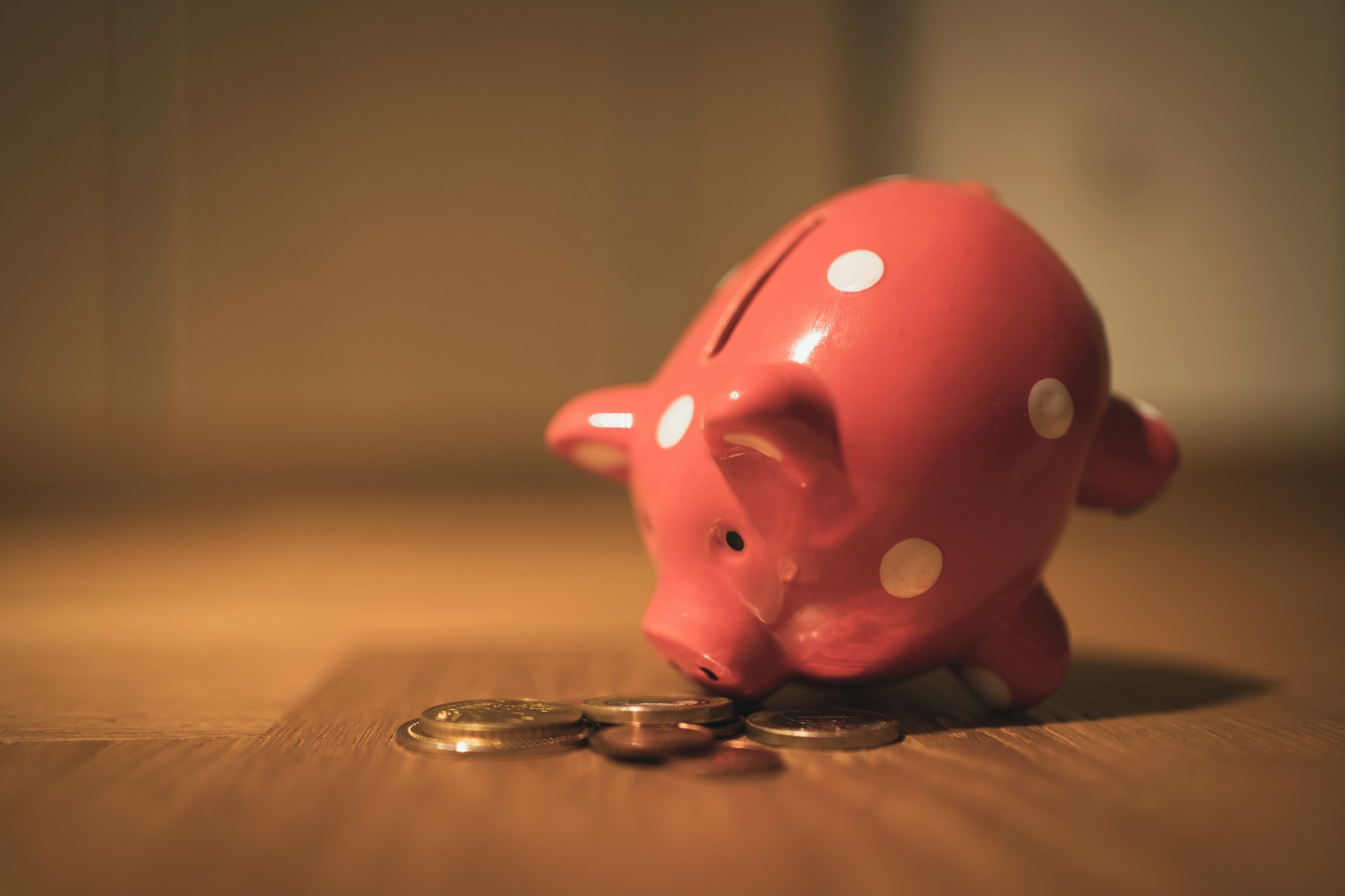 Pink piggy bank tipped over beside coins on a wooden floor