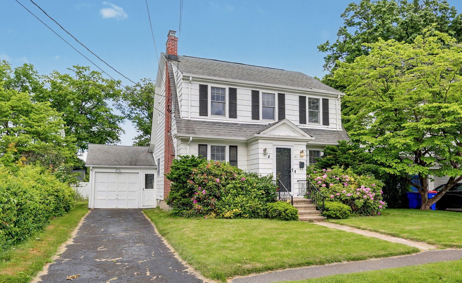 White gambrel-roofed Colonial with black shutters, a brick chimney and flowering shrubs, with a detached garage at the end of the driveway