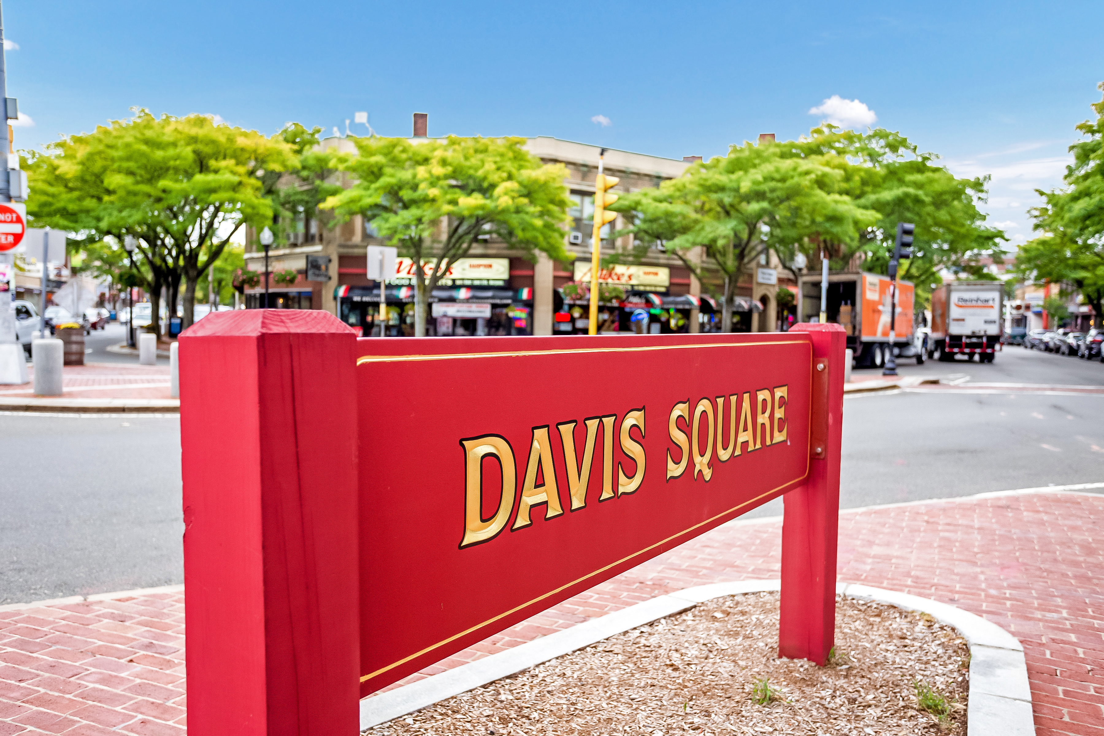 Red Davis Square sign in Somerville, Massachusetts with shops, trees, and neighborhood streets in the background.