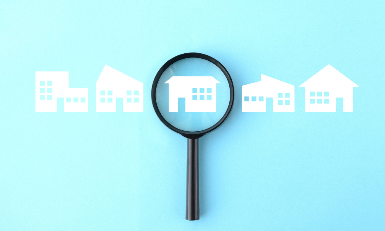 Magnifying glass highlighting a house icon among several home symbols on a light blue background, representing a focused home search.