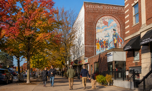 Autumn street scene in Union Square, Somerville, Massachusetts, with colorful foliage, pedestrians, local storefronts, and the historic Somerville mural.